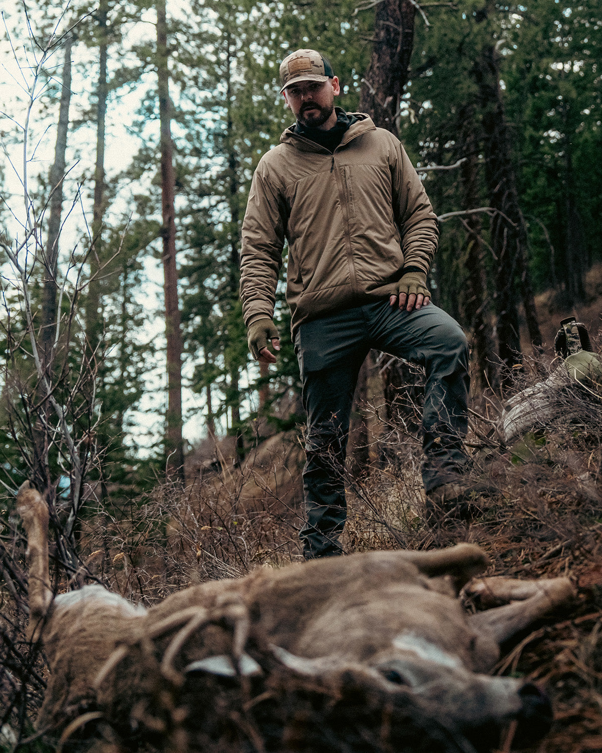 Ein Mann in Outdoor-Ausrüstung, der die EBERLESTOCK Alturas Hooded Jacket - Gunmetal trägt, steht in einem Wald neben einem großen Hirsch. Bäume und Laub umgeben sie und unterstreichen den Erfolg seines Jagdausflugs.