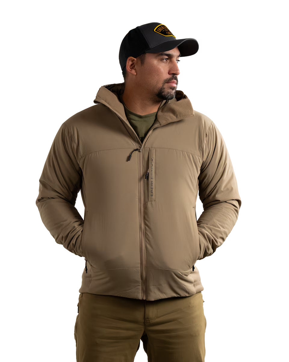 Ein Mann, der eine EBERLESTOCK Alturas Hooded Jacket in Dry Earth, ein olivgrünes Hemd und eine schwarze Mütze mit gelbem Aufnäher trägt, steht mit den Händen in den Taschen und blickt zur Seite vor einem weißen Hintergrund.