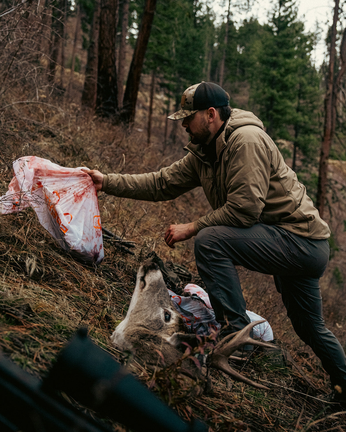 Ein Mann, der eine EBERLESTOCK Alturas Hooded Jacket - Dry Earth trägt, kniet mit einer blutigen Plastiktüte neben den Überresten eines Rehs auf einem bewaldeten Hügel, umgeben von Bäumen und trockenem Gestrüpp.