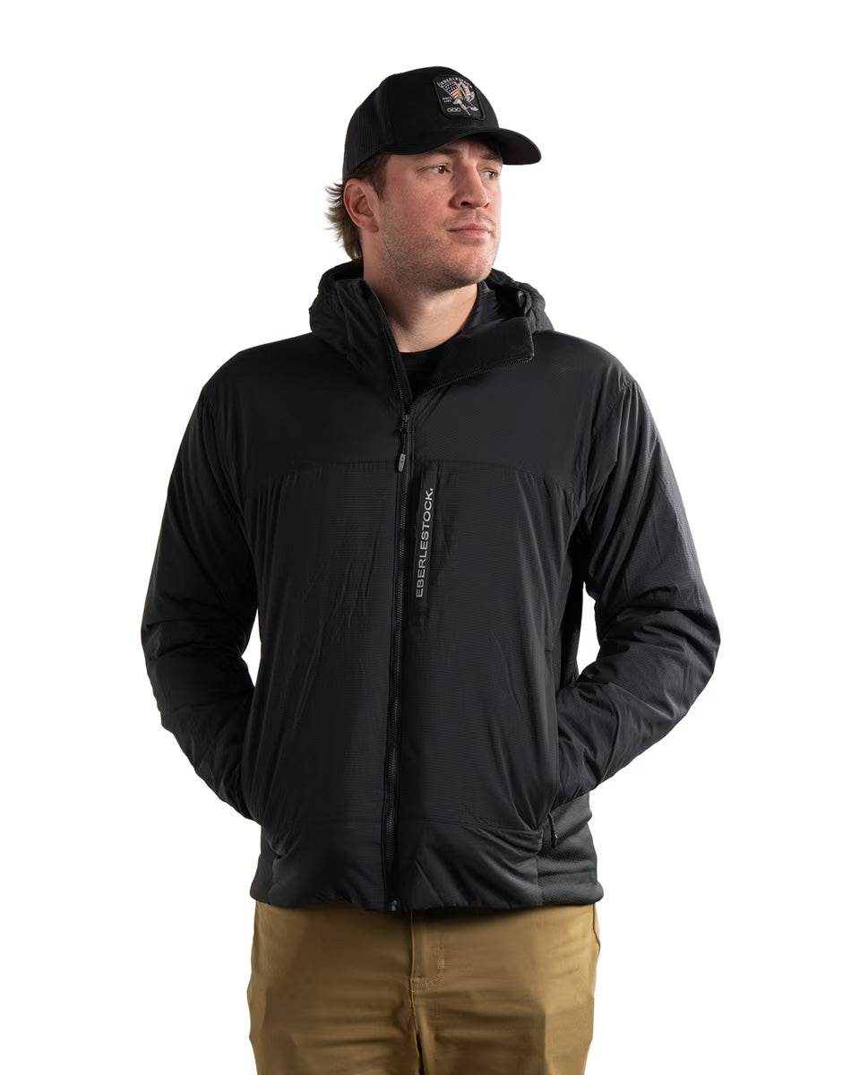Ein Mann steht mit den Händen in den Taschen, trägt die EBERLESTOCK Alturas Hooded Jacket - Black mit Frontreißverschluss, eine hellbraune Hose und eine schwarze Baseballkappe und blickt zur Seite vor einem schlichten weißen Hintergrund.