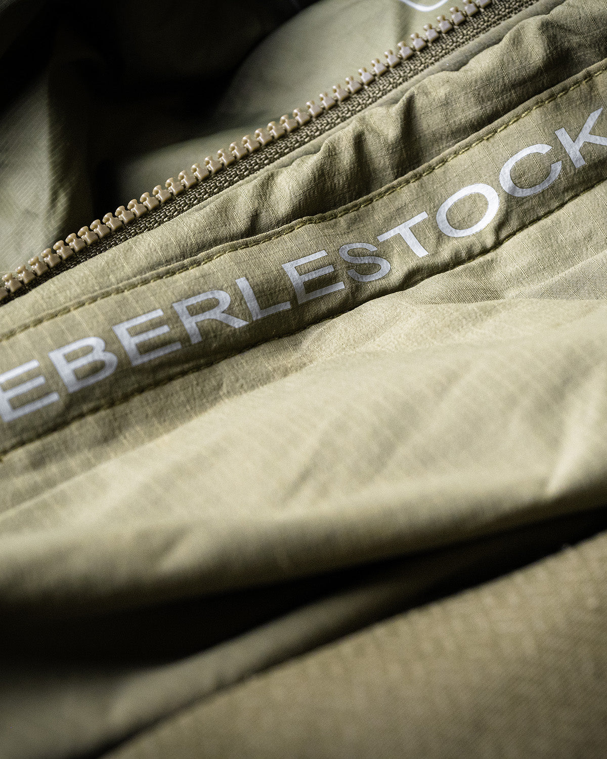 Nahaufnahme des schwarzen Stoffs einer Eberlestock Alturas Hooded Jacket von EBERLESTOCK, mit dem teilweise sichtbaren Markennamen neben einem metallischen Reißverschluss - ein Detail, das diese hochwertige Outdoor-Ausrüstung auszeichnet.