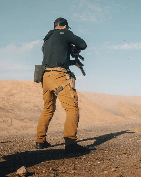 Eine Person mit schwarzem Kapuzenpulli, Mütze und EBERLESTOCK Deadwood Pant - Fall Green zielt mit einem Gewehr auf ein wüstenähnliches Gebiet im Freien und wirft einen langen Schatten auf den Boden unter einem teilweise bewölkten Himmel.