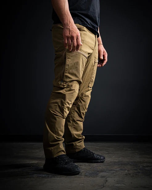 Eine Person steht auf einem dunklen Betonboden vor einem dunklen Hintergrund. Sie trägt eine EBERLESTOCK Deadwood Pant - Coyote Brown Cargohose, ein schwarzes T-Shirt und schwarze Turnschuhe, die Arme entspannt an der Seite.