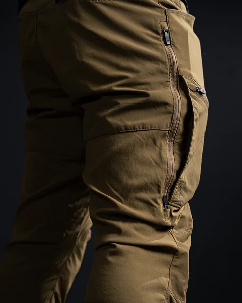 Nahaufnahme einer Person, die die EBERLESTOCK Deadwood Pant - Coyote Brown trägt, eine wasserabweisende Outdoor-Hose aus Stretch-Nylon mit mehreren Reißverschlüssen und Taschen, von hinten und von der Seite vor einem dunklen Hintergrund gezeigt.