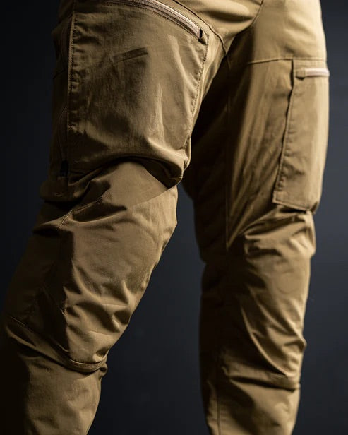 Nahaufnahme einer Person, die die EBERLESTOCK Deadwood Pant - Coyote Brown trägt, eine wasserabweisende Outdoor-Hose mit mehreren Taschen und sichtbaren Nähten, vor einem dunklen Hintergrund.