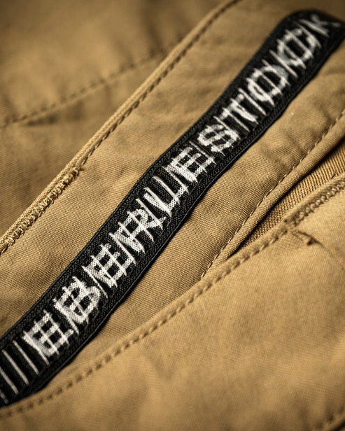 Nahaufnahme der koyotenbraunen Eberlestock Deadwood Pant von EBERLESTOCK mit schwarz-weißem Webetikett und stilisiertem Branding im Bereich des Reißverschlusses.