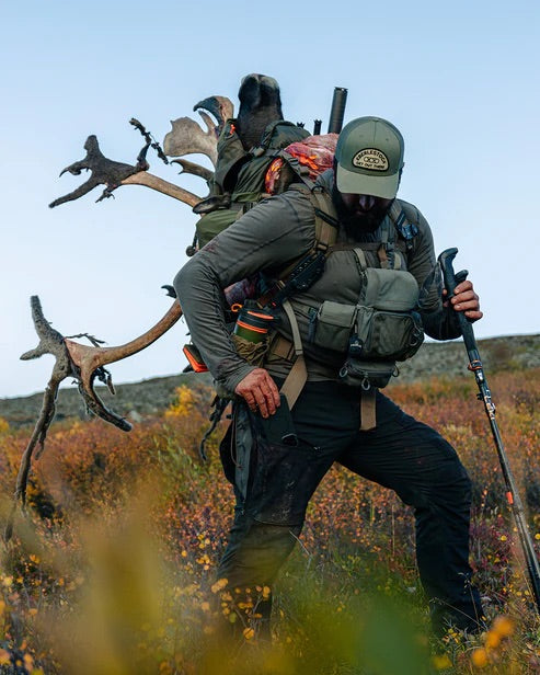 Ein bärtiger Mann in Outdoor-Ausrüstung und mit Mütze wandert durch das Unterholz. Er trägt die EBERLESTOCK Deadwood Pant - Coyote Brown, ideal für den Trail. Er trägt einen großen Rucksack mit Campingausrüstung und Hirschgeweih und benutzt Trekkingstöcke zur Unterstützung.
