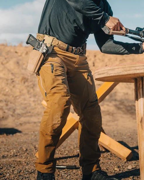 Eine Person in EBERLESTOCK Deadwood Pant - Coyote Brown und einem schwarzen langärmeligen Hemd steht im Freien an einem Holztisch und hält ein Gewehr mit einer Pistole im Holster am Gürtel in einer trockenen, schmutzigen Landschaft.