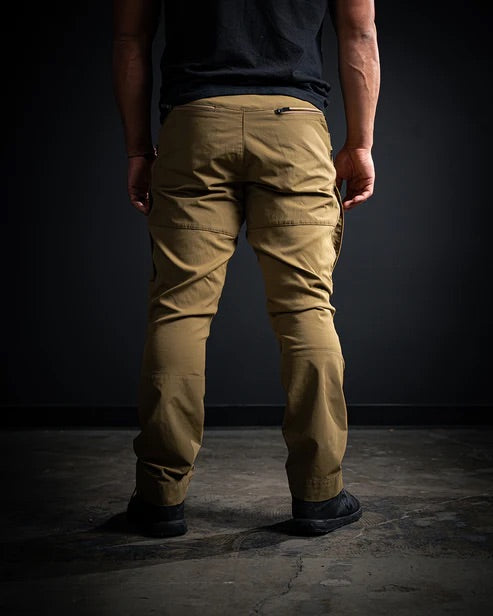 Eine Person steht, von der Kamera abgewandt, mit einem schwarzen T-Shirt, einer schwarzen EBERLESTOCK Deadwood Pant und schwarzen Schuhen auf einem dunklen Betonboden mit schwarzem Hintergrund.