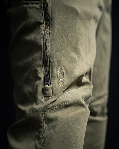 Nahaufnahme der EBERLESTOCK Camas Pro Pant - Fall Green, einer taktischen Hose mit einer Reißverschlusstasche und einem kleinen gestickten Logo mit drei ineinandergreifenden Ringen auf dem Oberschenkel.