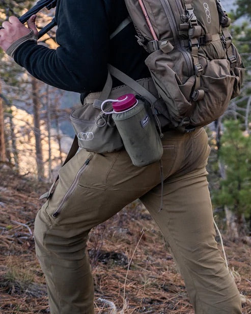 In der EBERLESTOCK Camas Pro Pant in Fall Green und einer schwarzen Jacke erkundet ein Wanderer mit einem großen braunen Rucksack und einer Wasserflasche die Natur zwischen Kiefern und trockenem Gras.