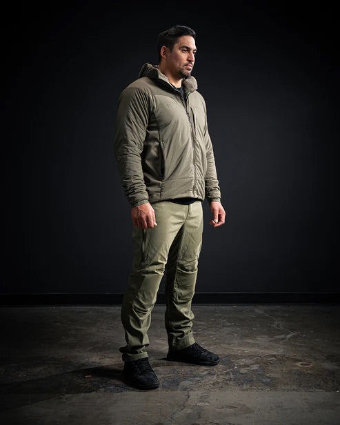 Ein Mann steht in einem Innenraum vor einem dunklen Hintergrund. Er trägt eine olivgrüne Kapuzenjacke, eine EBERLESTOCK Camas Pro Pant - Black und schwarze Schuhe, die Arme liegen entspannt an seinen Seiten.