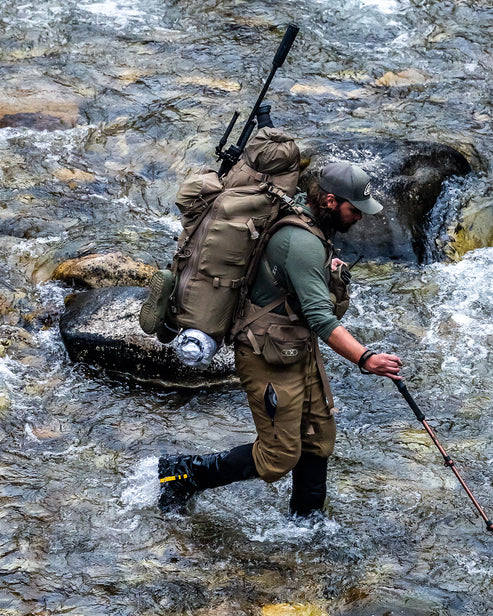 Ein bärtiger Jäger watet durch einen felsigen Gebirgsbach, trägt einen taktischen Rucksack und ein Gewehr, benutzt einen Trekkingstock, trägt robuste Stiefel und die EBERLESTOCK Camas Pro Pant - Black.