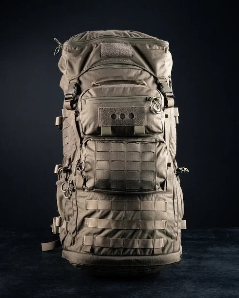Der EBERLESTOCK Mission Ruck, ein großer hellbrauner Rucksack mit mehreren Fächern, Riemen, Schnallen und MOLLE-Gewebe, steht aufrecht vor einem dunklen Hintergrund.