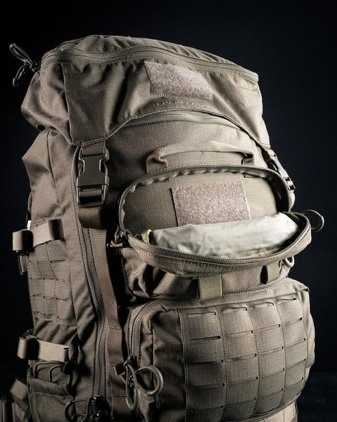 Eine Nahaufnahme des EBERLESTOCK Mission Rucksacks zeigt seine hellbraune Farbe, mehrere Fächer und Riemen sowie eine teilweise geöffnete Tasche, die ein helles Inneres offenbart, alles vor einem dunklen Hintergrund.