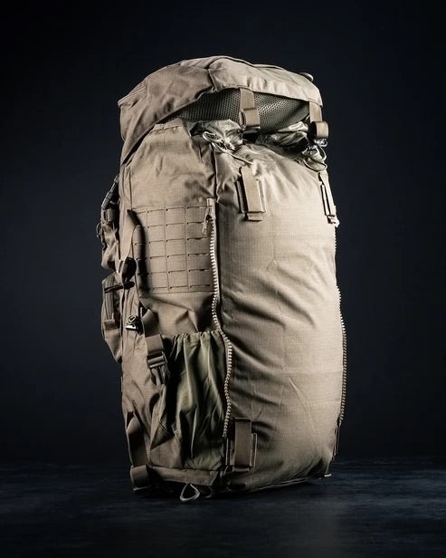 Der EBERLESTOCK Mission Ruck, ein großer hellbrauner taktischer Rucksack mit zahlreichen Riemen, Reißverschlüssen und Taschen, steht aufrecht vor einem dunklen, minimalistischen Hintergrund.