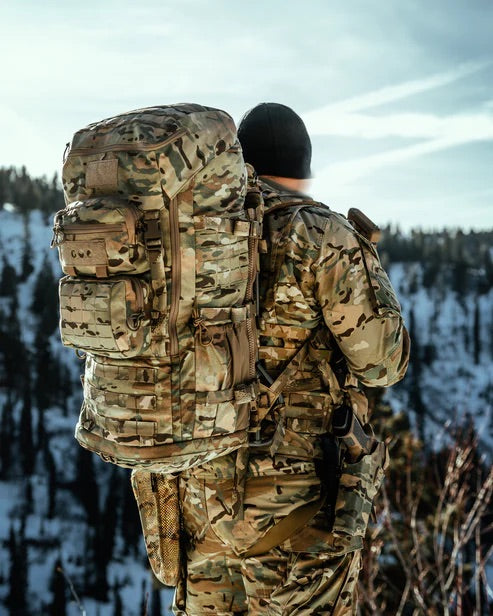 Eine in Tarnkleidung gekleidete Person mit einem EBERLESTOCK Mission Ruck steht im Freien und bewundert die schneebedeckten Berge unter einem teilweise bewölkten Himmel.