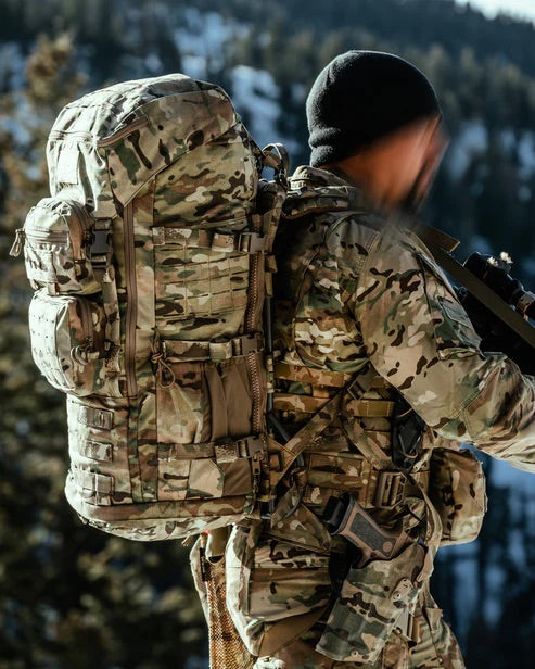 Eine Person in Tarnkleidung steht mit einem EBERLESTOCK Mission Ruck Rucksack und einem Gewehr in einem bewaldeten Berggebiet; ihr Gesicht und ihre Hände zeigen verschwommene Bewegungen.