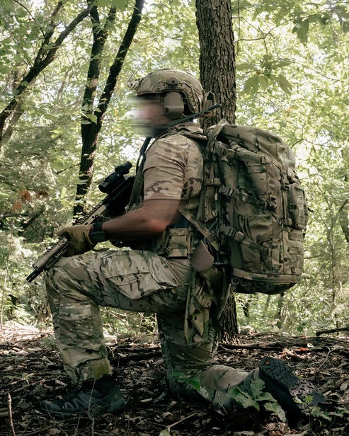Ein Soldat in Tarnkleidung und Helm kniet in einem sonnenbeschienenen Wald mit einem Gewehr und einem EBERLESTOCK Mission Ruck. Sein Gesicht ist zur Anonymität unscharf.