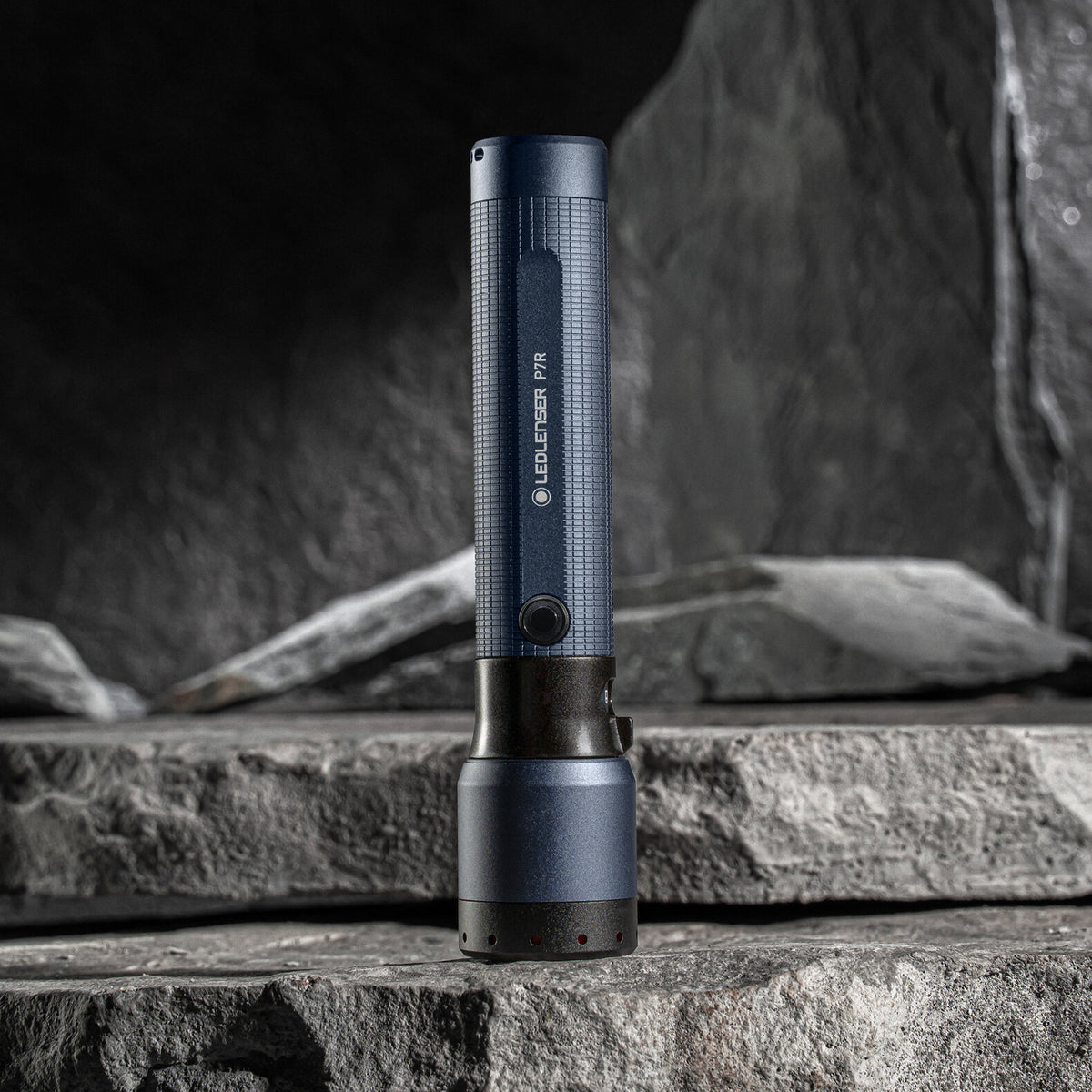 Die LEDLENSER Taschenlampe P7R - 25 Jahre Edition, in dunkelblau und schwarz, steht aufrecht auf gestapelten grauen Felsen. Diese besondere Taschenlampe hat einen strukturierten Griff, einen sichtbaren Knopf und liefert 2000 Lumen Helligkeit.