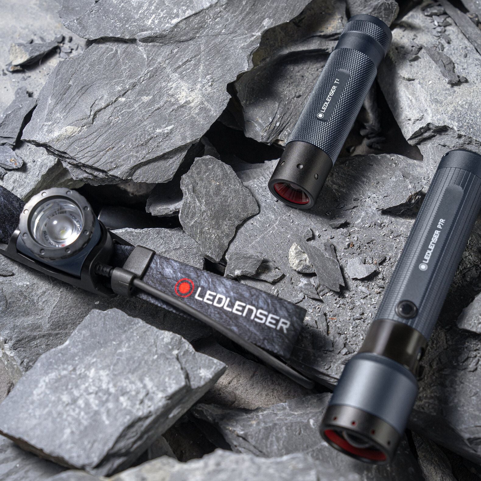 Drei LEDLENSER-Leuchten, darunter die Ledlenser Taschenlampe P7R - 25 Jahre Edition und eine Stirnlampe mit verstellbarem LEDLENSER-Band, ruhen auf grauen Felsen. Die schwarzen und grauen Geräte sind mit roten Akzenten versehen.
