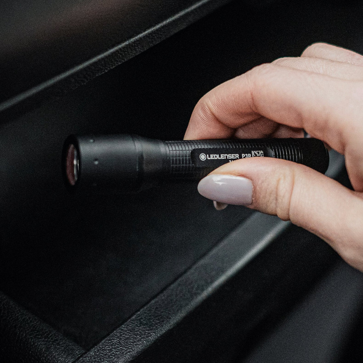 Eine Hand mit hellviolettem Nagellack hält eine kompakte LEDLENSER Taschenlampe P3R neben einem schwarzen Regal oder einer Schublade.