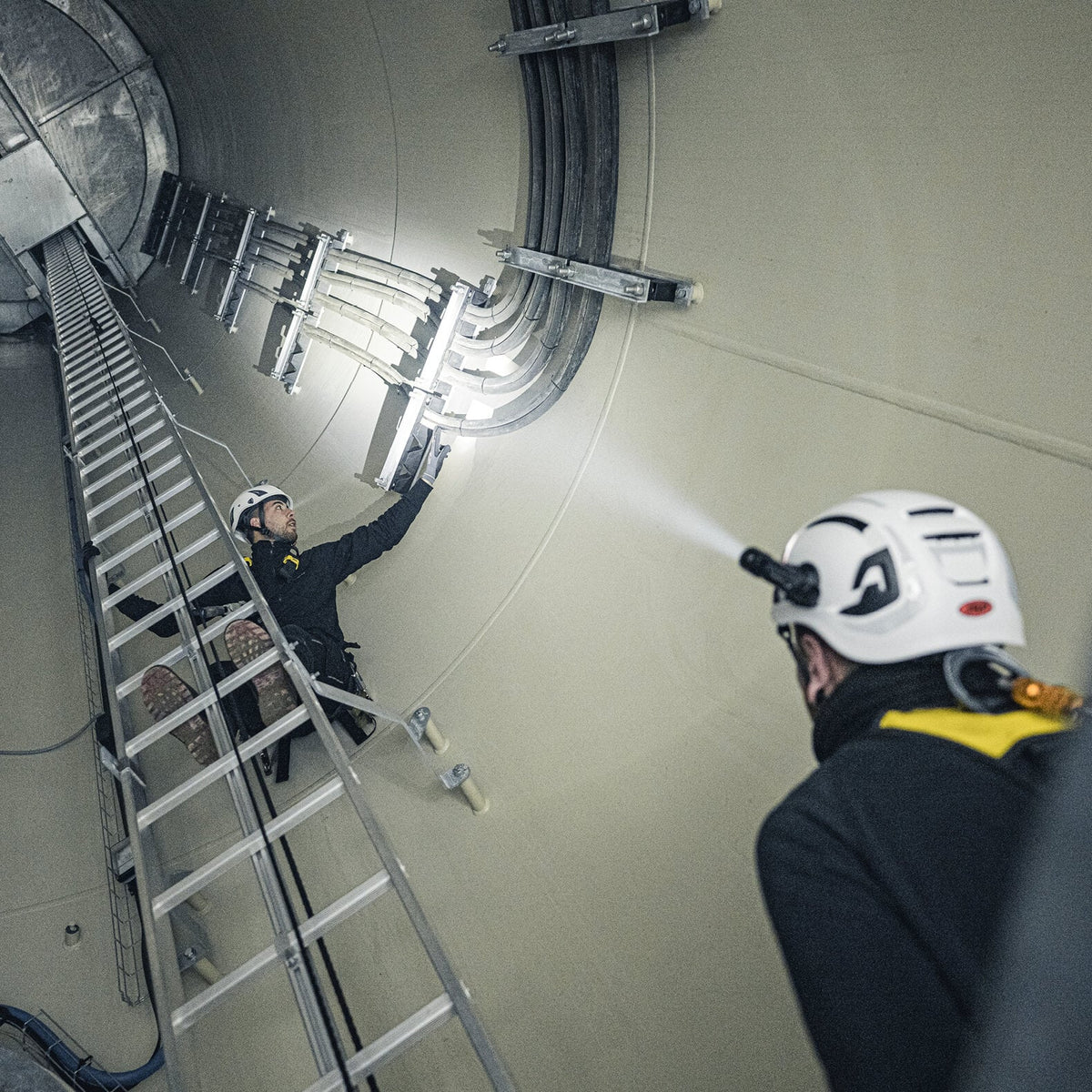 Zwei Arbeiter mit LEDLENSER P5R-Scheinwerfern inspizieren elektrische Kabel entlang der Wand in einem großen, zylindrischen Industrietunnel, wobei die Leitern vertikal aufgestellt sind.