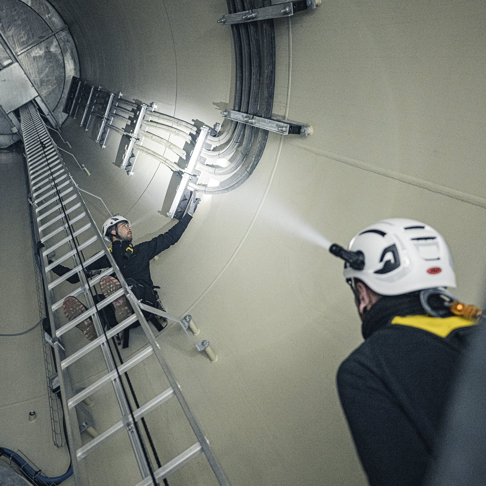 Zwei Arbeiter mit LEDLENSER P5R-Scheinwerfern inspizieren elektrische Kabel entlang der Wand in einem großen, zylindrischen Industrietunnel, wobei die Leitern vertikal aufgestellt sind.