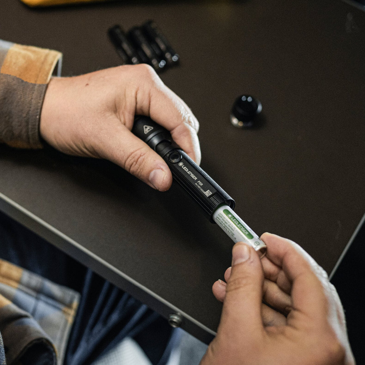 Jemand legt eine wiederaufladbare AA-Batterie in eine schwarze LEDLENSER Taschenlampe P5R ein, während er an einem Tisch steht, mit Ersatzbatterien und dem Batteriedeckel in der Nähe.