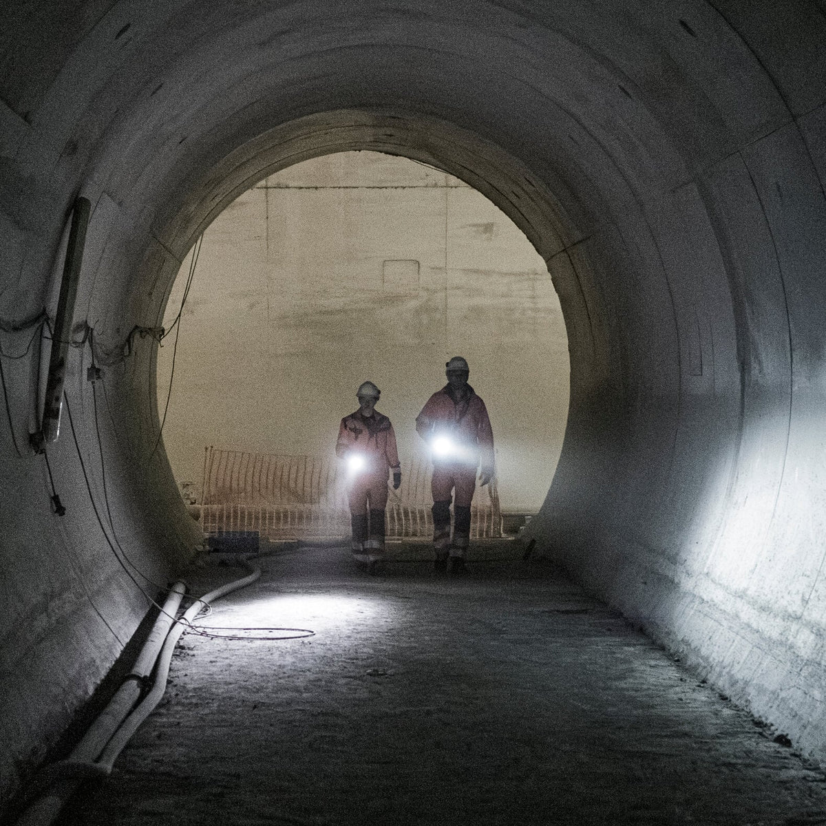 Zwei Bauarbeiter mit Helmen und Schutzwesten gehen durch einen großen, schwach beleuchteten Tunnel und halten dabei helle LEDLENSER Ledlenser Taschenlampen P7R Pro, die ideal für raue Umgebungen im Freien sind.