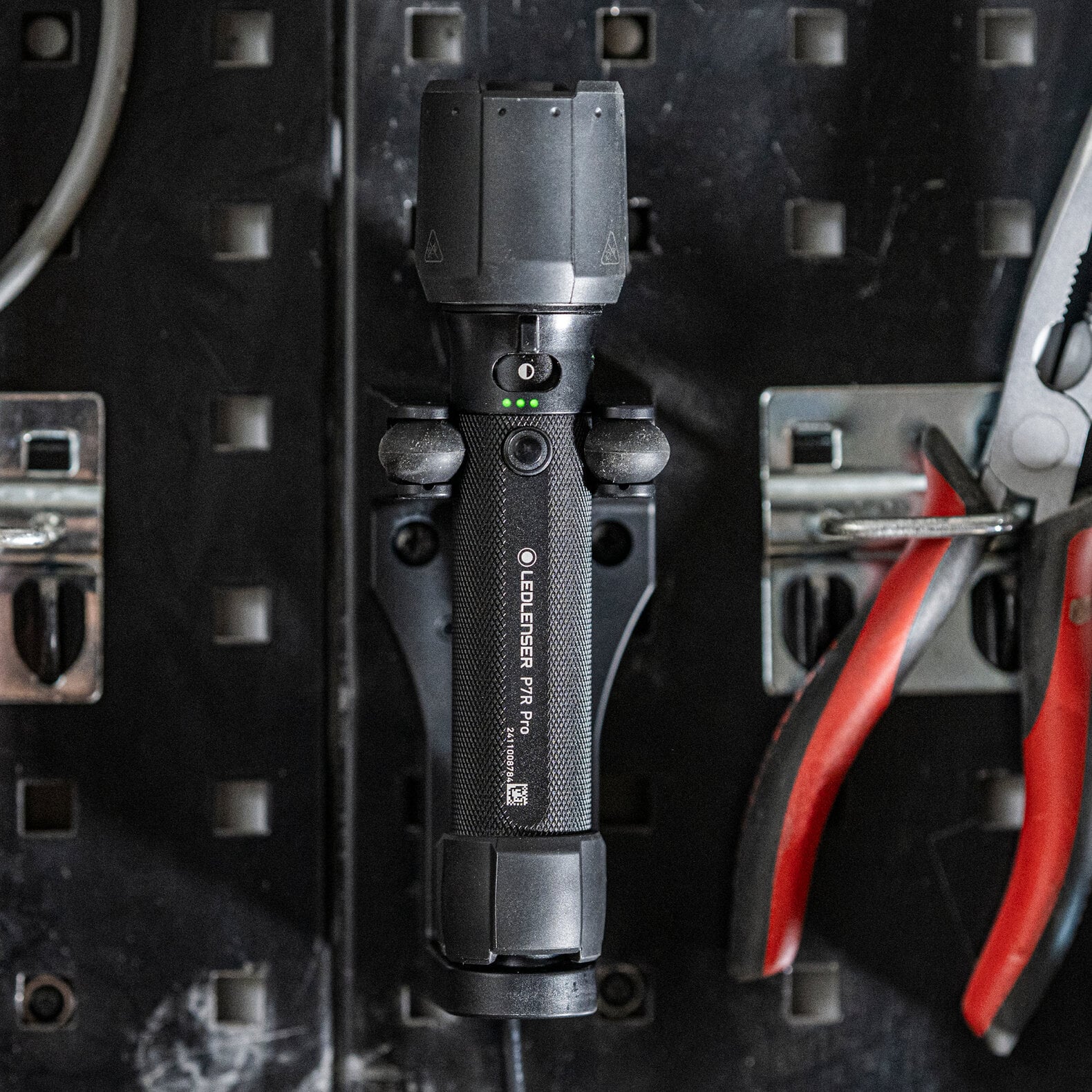 Eine schwarze LEDLENSER Taschenlampe P7R Pro ist senkrecht auf einem Lochblechbrett montiert, flankiert von einer rot lackierten Zange und einem grauen Kabel.
