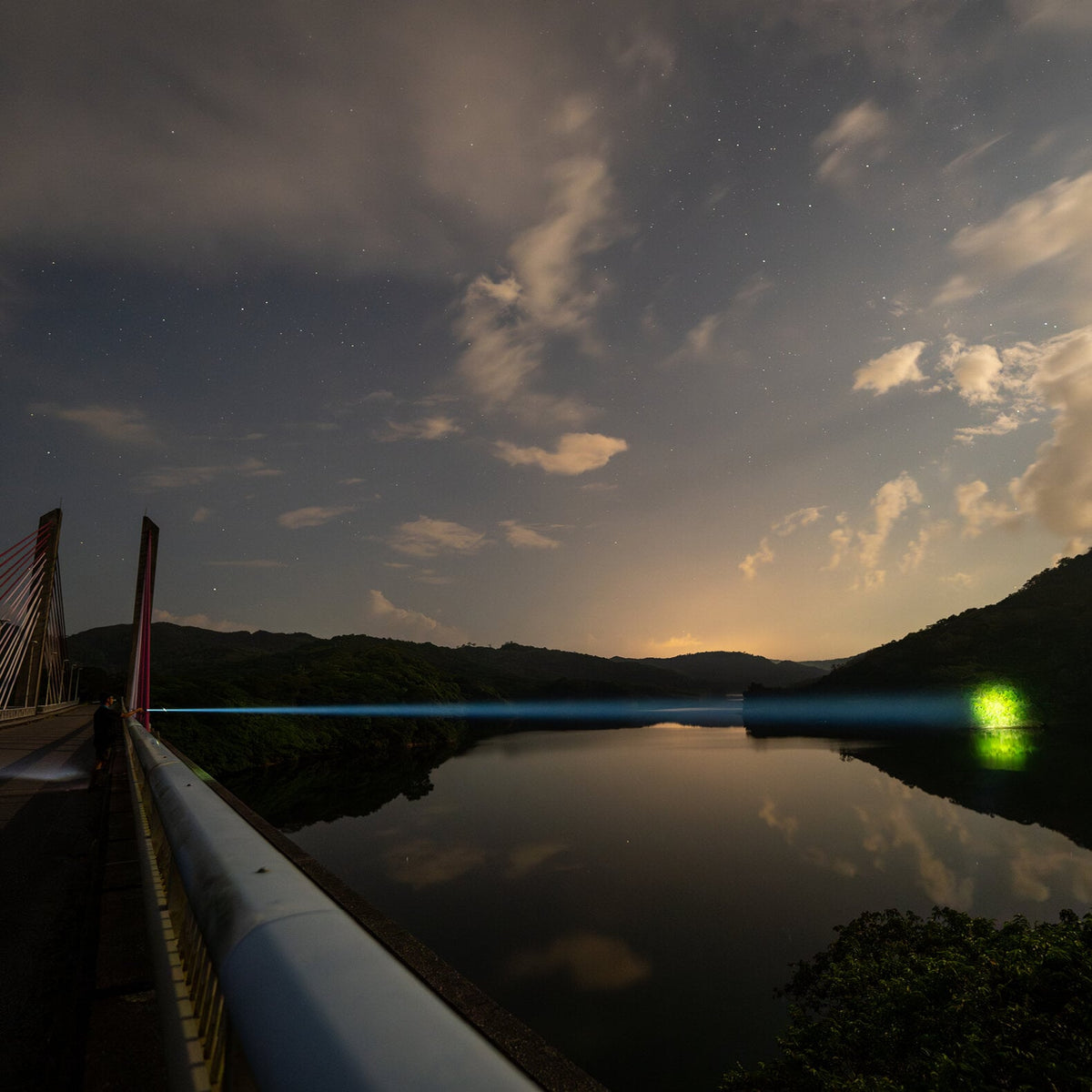 Ein nächtlicher Blick von einer Schrägseilbrücke über einen ruhigen Fluss, wobei der blaue Laserstrahl der LEDLENSER Taschenlampe P9R Core LEP das Wasser in Richtung eines grünen Lichts überquert, unter einem orangen Himmel über bewaldeten Hügeln.