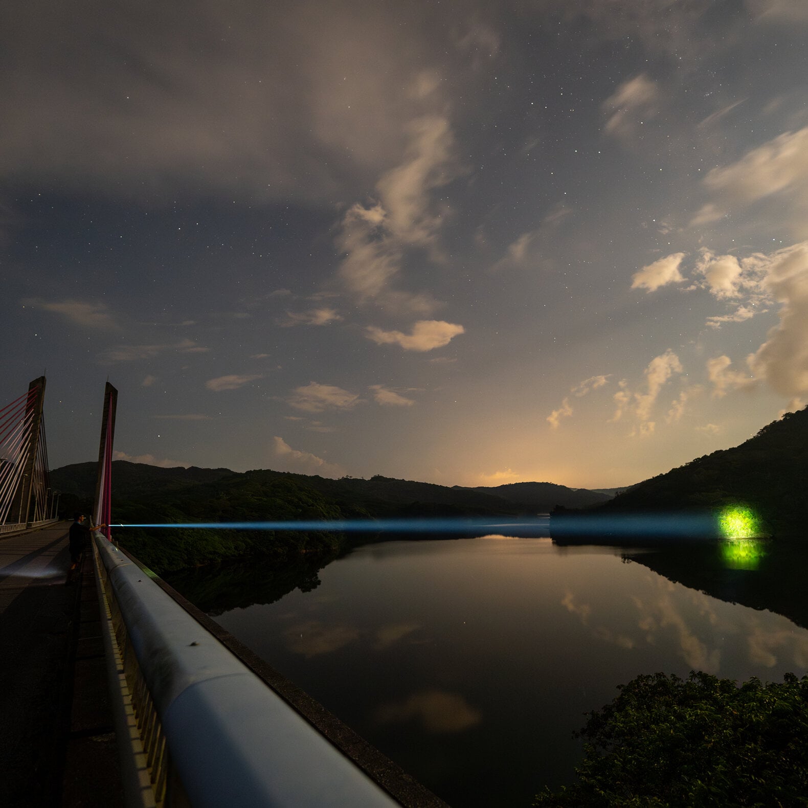 Ein nächtlicher Blick von einer Schrägseilbrücke über einen ruhigen Fluss, wobei der blaue Laserstrahl der LEDLENSER Taschenlampe P9R Core LEP das Wasser in Richtung eines grünen Lichts überquert, unter einem orangen Himmel über bewaldeten Hügeln.