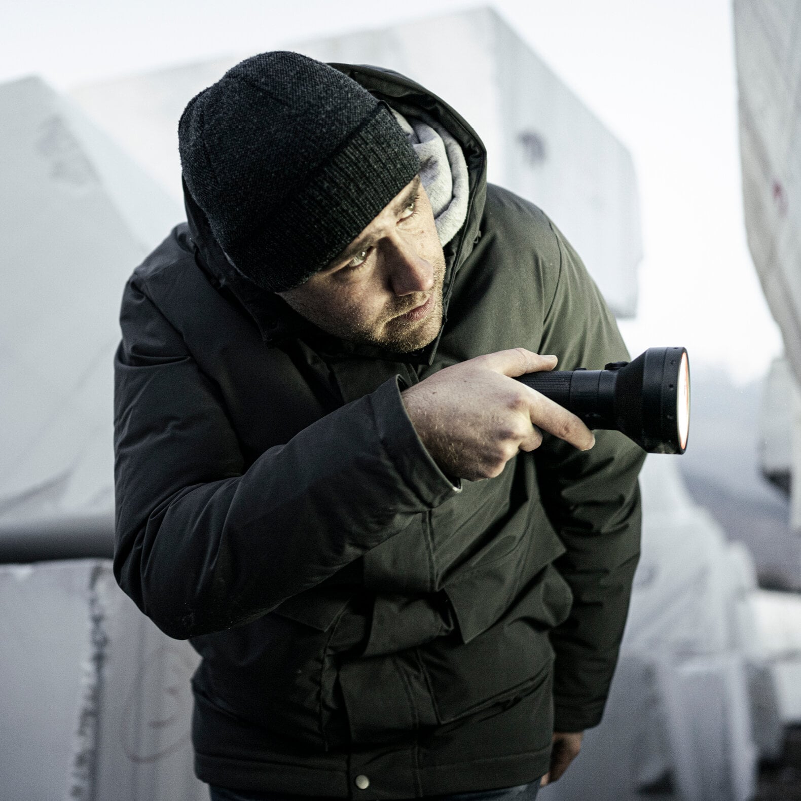 Ein Mann mit schwarzer Mütze und Winterjacke hält die LEDLENSER Ledlenser Taschenlampe P18R in der Hand, beugt sich vor und blickt nach draußen, umgeben von großen weißen rechteckigen Blöcken.