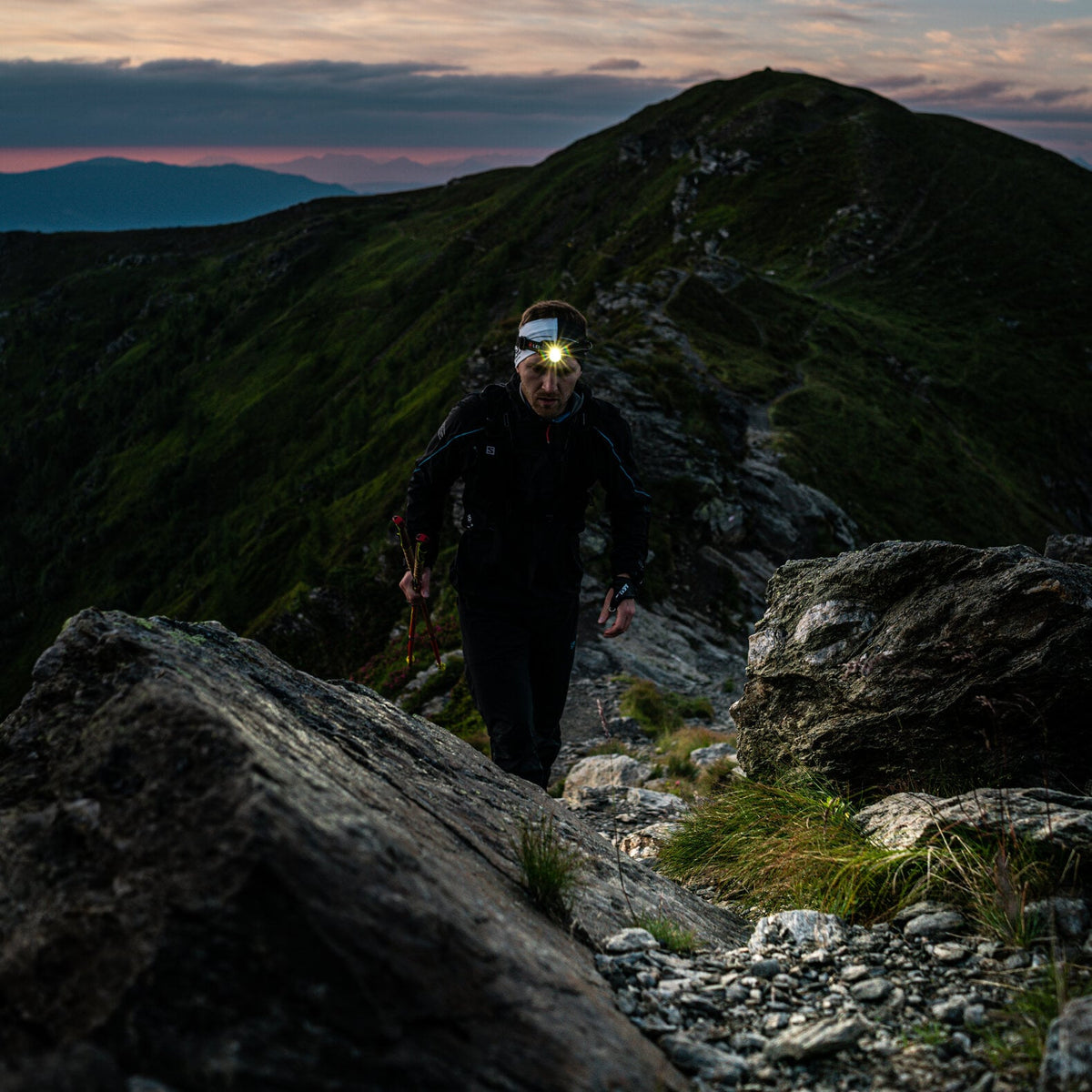Ein Wanderer, der eine LEDLENSER Stirnlampe H7R Core mit Advanced Focus System trägt, steigt in der Dämmerung einen felsigen Bergpfad hinauf, mit grünen Hängen und dramatischen Wolken im Hintergrund.