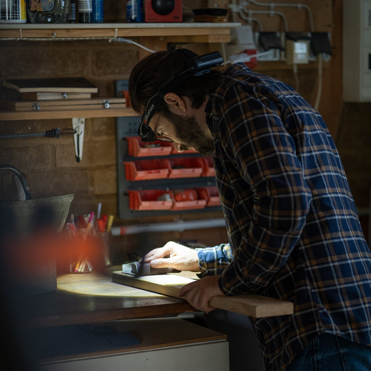 Ein Mann, der eine LEDLENSER Ledlenser Stirnlampe H7R Core mit Advanced Focus System und ein kariertes Hemd trägt, arbeitet in einer schwach beleuchteten Werkstatt sorgfältig an Holz, umgeben von Werkzeugen und Vorräten in Regalen.