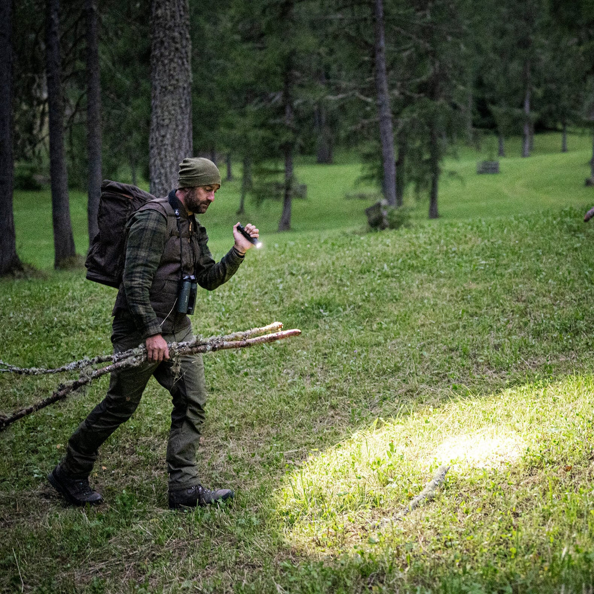 Ein Mann in Outdoor-Ausrüstung geht durch einen grasbewachsenen Wald, in der Hand Äste und eine LEDLENSER Ledlenser Taschenlampe TT3R, deren Lichtstrahl einen hellen Fleck auf dem Boden beleuchtet.