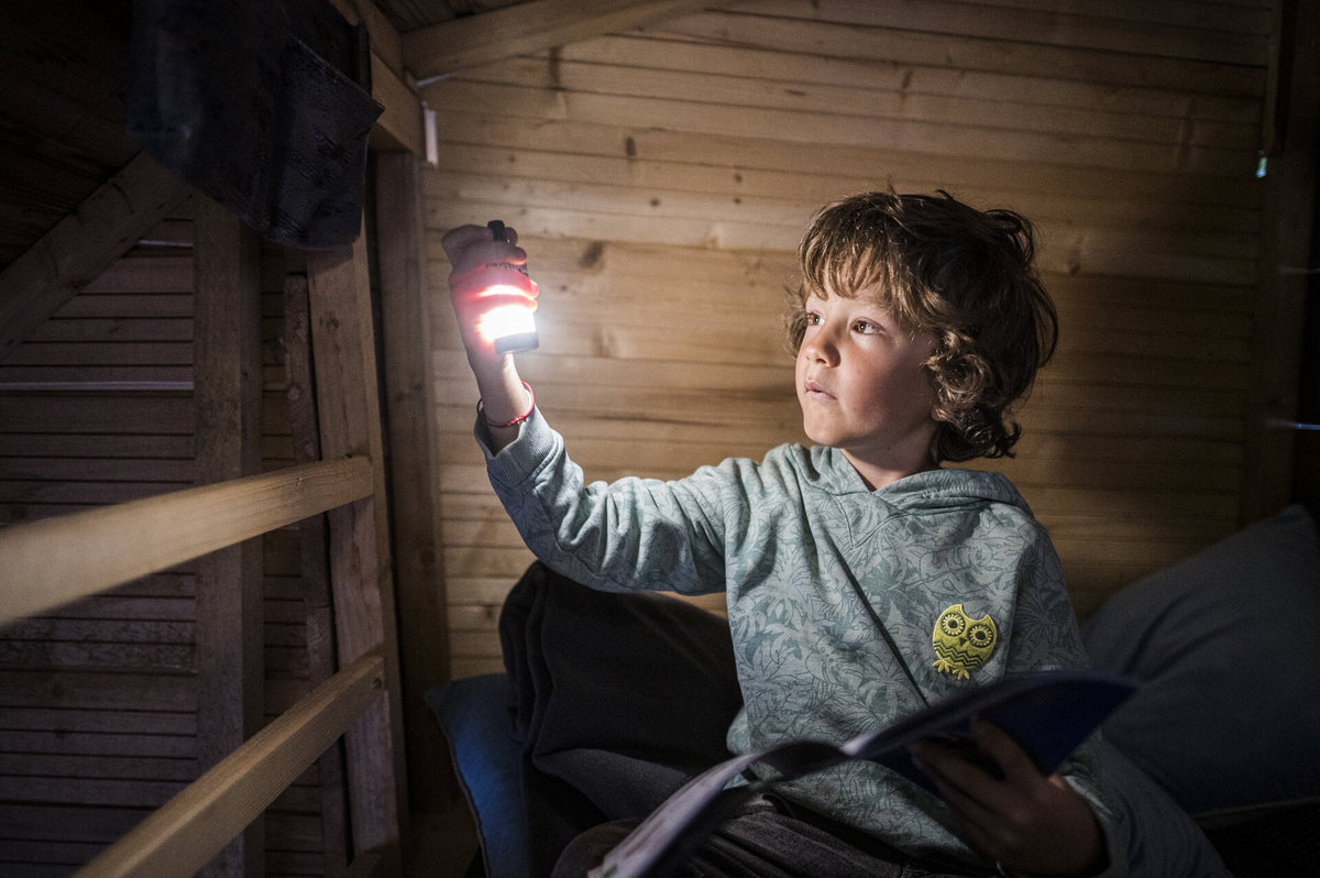 Ein kleines Kind sitzt in einem Holzzimmer und hält eine LEDLENSER Ledlenser ML4 Outdoor Camping Lampe in der Hand. Ihr weiches Licht strahlt nach oben und beleuchtet das Gesicht des Kindes und die holzgetäfelten Wände für eine gemütliche Leseatmosphäre.