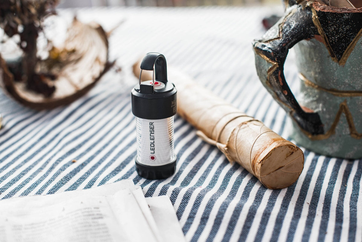 Eine kompakte LEDLENSER ML4 Outdoor-Campinglampe mit Dual-Power-Technologie steht auf einem blau-weiß gestreiften Tuch neben einem doppelt verpackten Brötchen, einem dekorativen Becher, einem Stück Zeitung und einer getrockneten Pflanze.