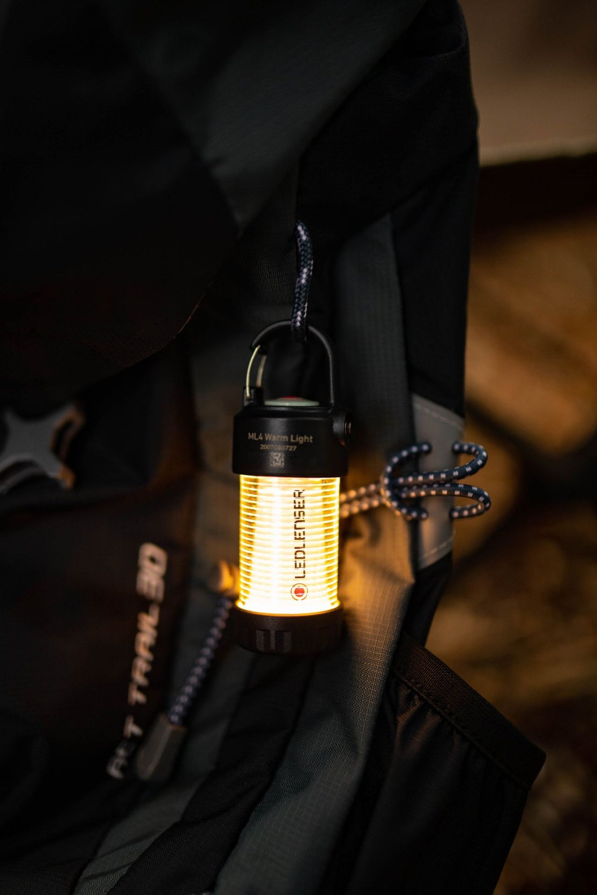 Eine kompakte LEDLENSER ML4 WL Outdoor Camping Lampe mit warmweißem Licht hängt an einer Schlaufe an einem dunklen Rucksack und beleuchtet sanft den Stoff und das Seil in der Nähe. Die Lampe ist mit "HL4 Warm Light" beschriftet und mit der Marke LEDLENSER versehen.