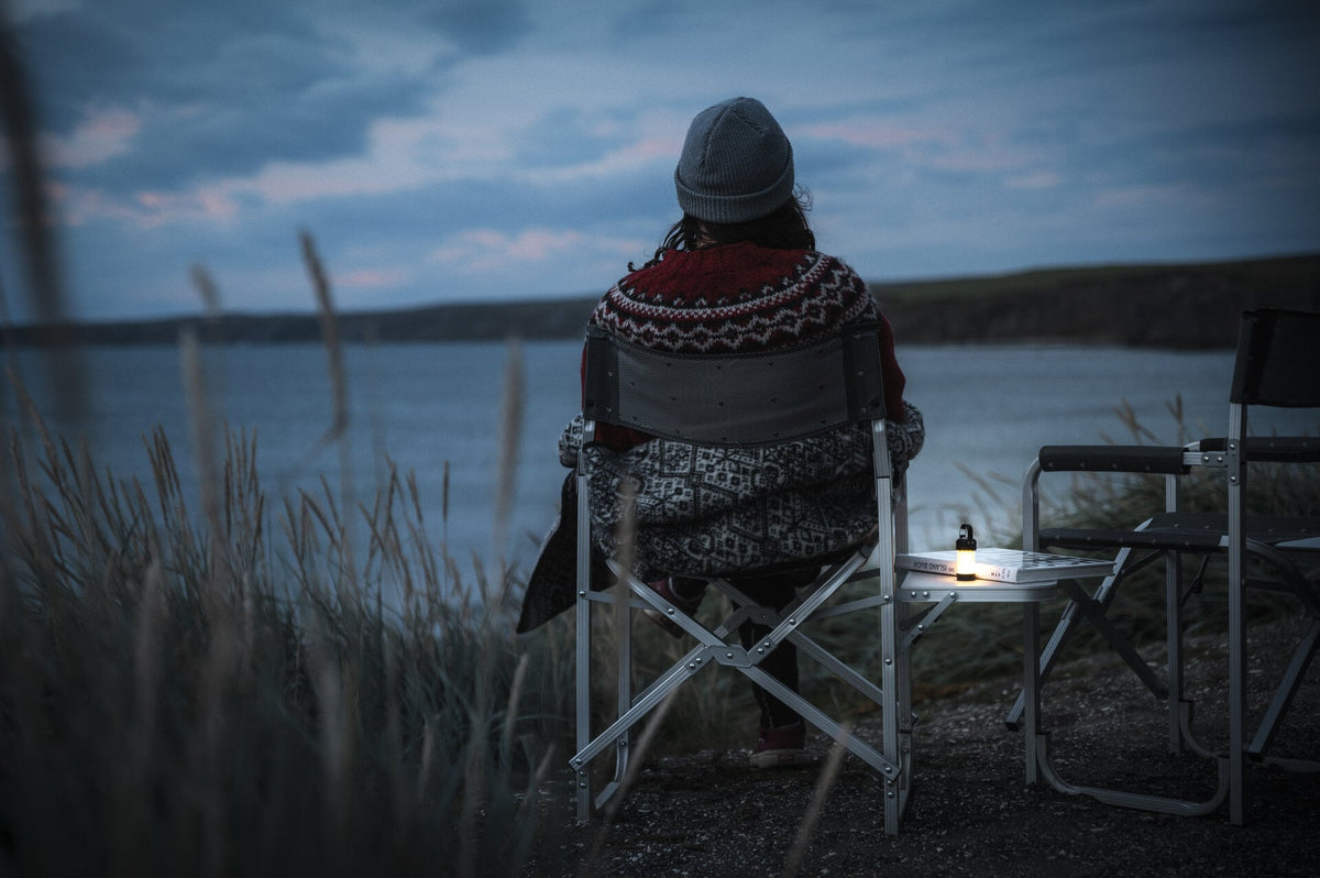 Eine Person, die einen Strickpullover und eine Mütze trägt, sitzt in der Abenddämmerung auf einem Klappstuhl am Wasser. Eine LEDLENSER Ledlenser ML4 WL Outdoor Camping Lampe und ein Buch sind auf einem anderen Stuhl platziert, mit hohem Gras im friedlichen Vordergund.