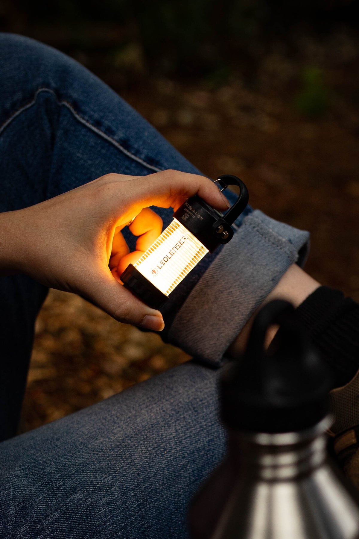 Eine Person in Jeans hält eine leuchtende LEDLENSER ML4 WL Outdoor Camping Lampe in der Nähe ihres Knies. Im Vordergrund ist eine Wasserflasche aus Edelstahl zu sehen, im Hintergrund eine unscharfe Naturkulisse.