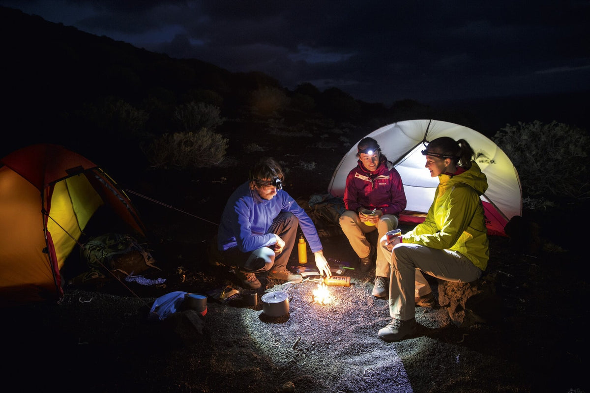 Drei Personen sitzen nachts an einem kleinen Lagerfeuer zwischen zwei Zelten, tragen Outdoor-Jacken und eine LEDLENSER Stirnlampe H14R.2 mit Advanced Focus System und Akku und teilen im sanften Schein warme Getränke und Speisen.