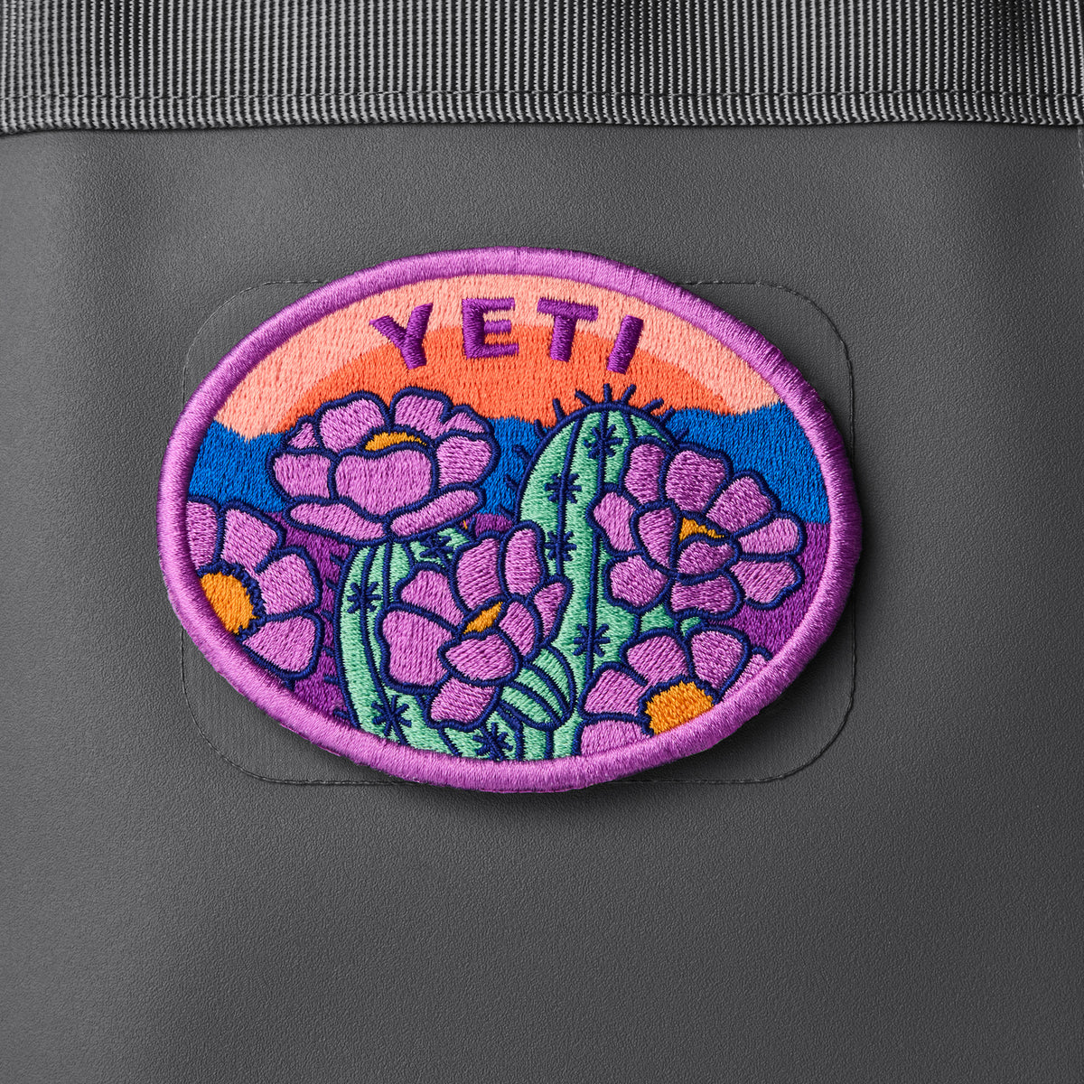 Das YETI® Full Bloom Patch von YETI zeigt eine farbenfrohe Wüstenszene - lilafarbene Sukkulenten, einen grünen Kaktus, orangefarbene Akzente und einen farbenfrohen Sonnenuntergang -, die mit einem Klettverschluss an Ihrer grauen Tasche befestigt wird; perfekt für Ihre Outdoor-Ausrüstung.