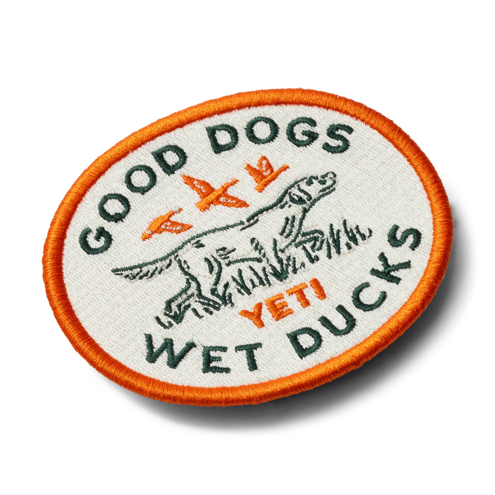 Der YETI® Good Dogs Patch ist ein kreisförmig gestickter Aufnäher mit orangefarbenem Rand, der einen Hund, zwei fliegende Enten und den Schriftzug "GOOD DOGS WET DUCKS YETI" zeigt - ideal für Outdoor-Ausrüstung und Klettflächen.