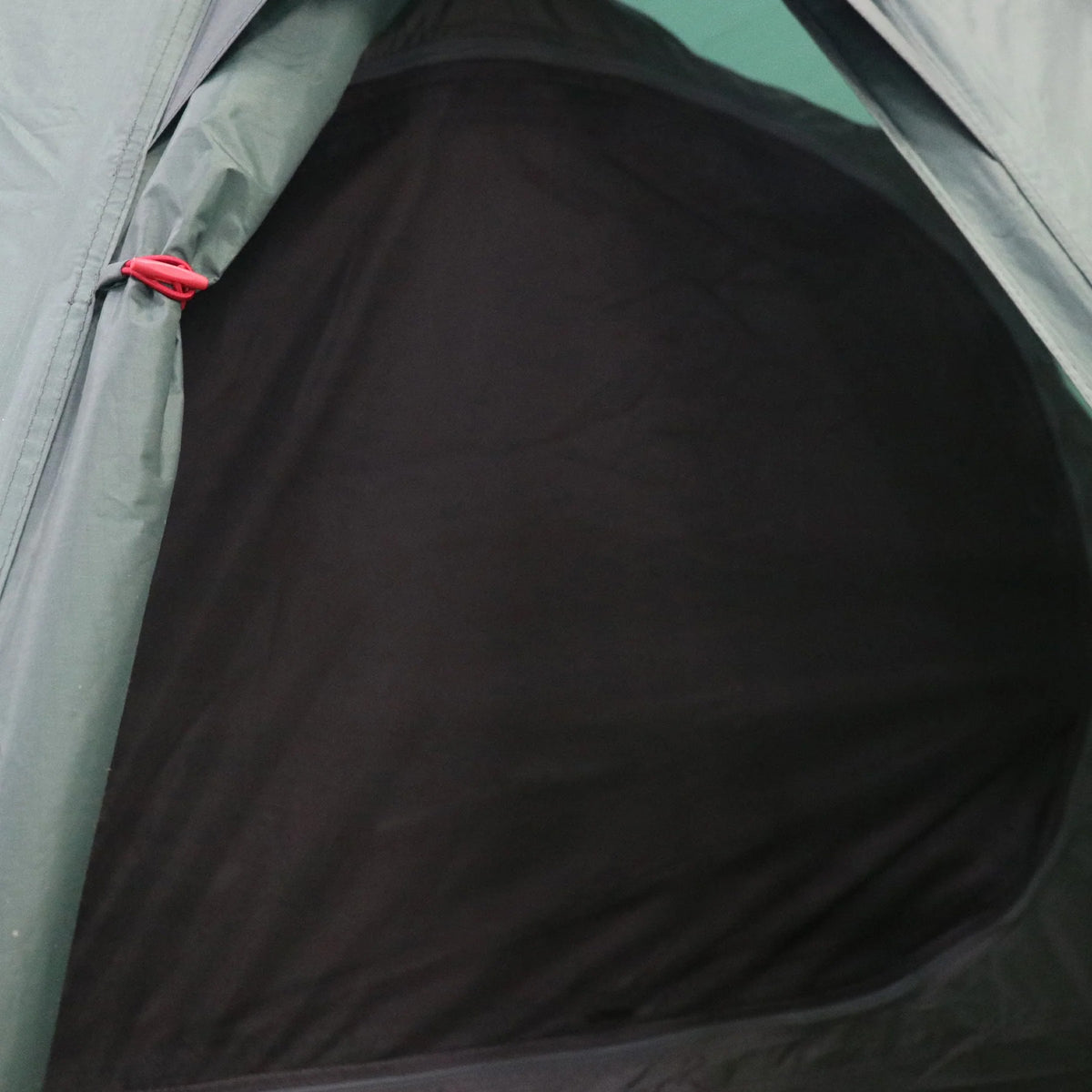 Nahaufnahme des Eingangs zu einem HIGHLANDER FORCES Highlander Respite 1 Personen Tunnelzelt - Pine Green. Die Mesh-Reißverschlusstür wird mit einem roten Clip geschlossen, und das schummrige Innere ist durch das Mesh sichtbar.