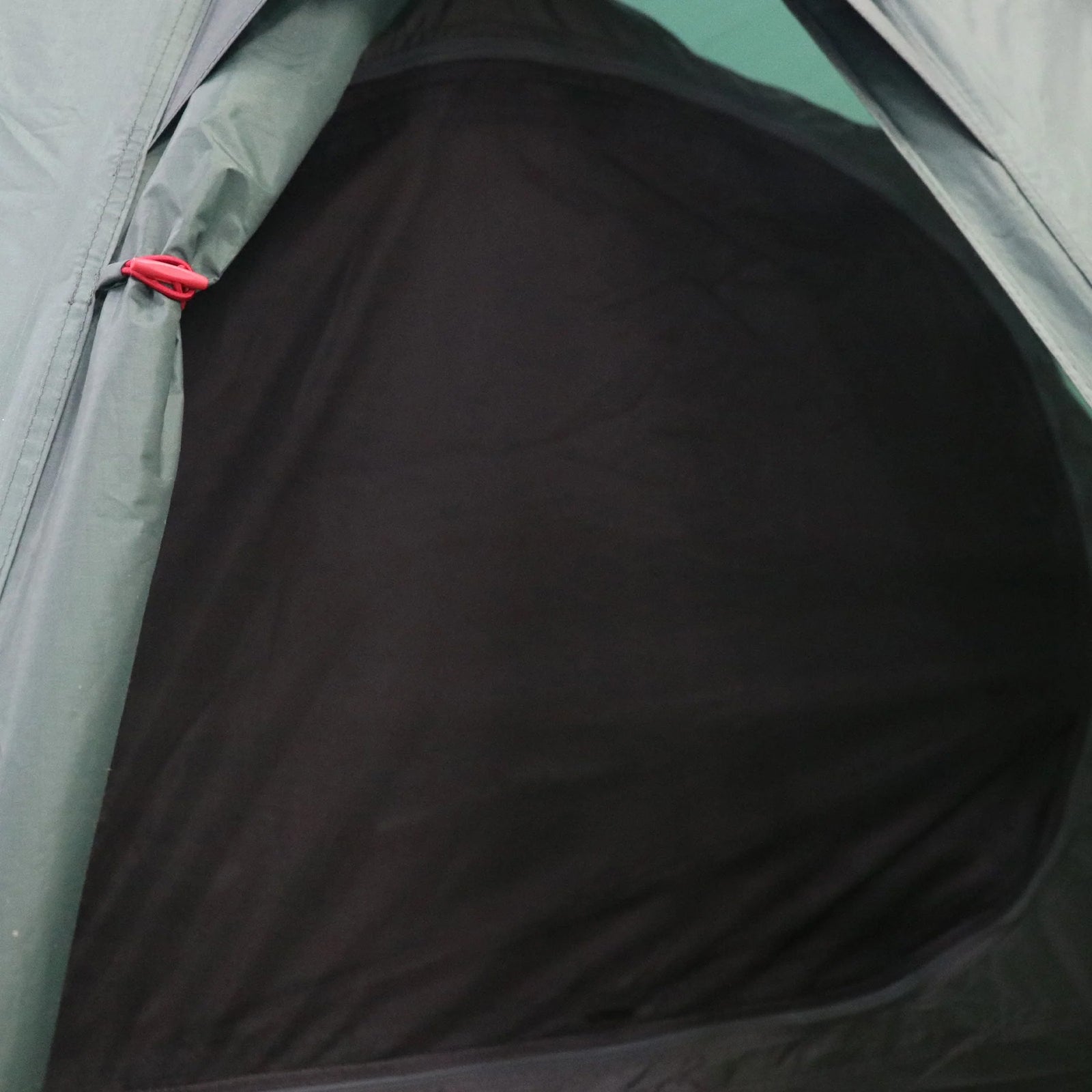 Nahaufnahme des Eingangs zu einem HIGHLANDER FORCES Highlander Respite 1 Personen Tunnelzelt - Pine Green. Die Mesh-Reißverschlusstür wird mit einem roten Clip geschlossen, und das schummrige Innere ist durch das Mesh sichtbar.