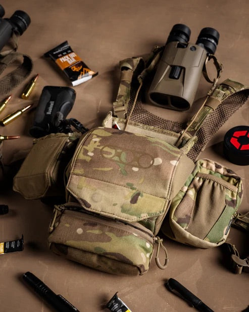 Ein tarnfarbenes taktisches Brustgeschirr mit mehreren Taschen ruht auf einer braunen Fläche, umgeben von Jagdausrüstung wie Ferngläsern, Gewehrmunition, Energieriegeln, schwarzen Schläuchen und einem EBERLESTOCK Recon Modular Bino Pack Large.