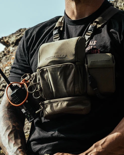 Draußen im Sonnenlicht zeigt eine Person in schwarzem Hemd und olivgrüner taktischer Brustausrüstung mit Taschen und Funkgerät ihr EBERLESTOCK Eberlestock Recon Modular Bino Pack Large als Teil ihrer Jagdausrüstung.