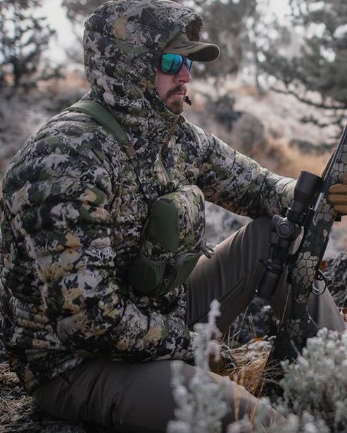 Eine Person in Tarnkleidung mit einem Gewehr mit Zielfernrohr sitzt im Freien inmitten von trockenem Gras und Felsen. Das EBERLESTOCK Recon Modular Bino Pack Large fügt sich nahtlos in die raue Landschaft ein und ergänzt die natürliche Umgebung.